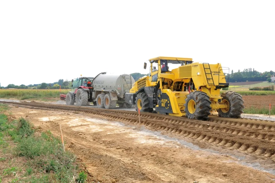 Derrière le malaxeur Bomag RS 650, un technicien vérifie visuellement l'homogénéité du mélange entre le matériau de l’ancienne chaussée et le liant hydraulique, et un autre mesure la teneur en eau du matériau traité.