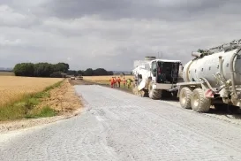 Vue générale du chantier avec, au premier plan, la machine de malaxage alimentée en eau par une citerne, et en arrière plan,  la niveleuse et l'atelier de compactage.